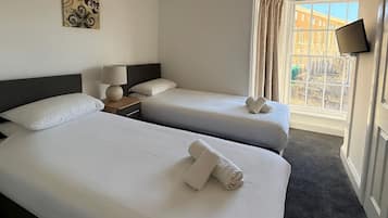 Habitación de hotel con dos camas, una mesita de noche con lámpara, un televisor de pantalla plana montado en la pared y una ventana con cortinas.