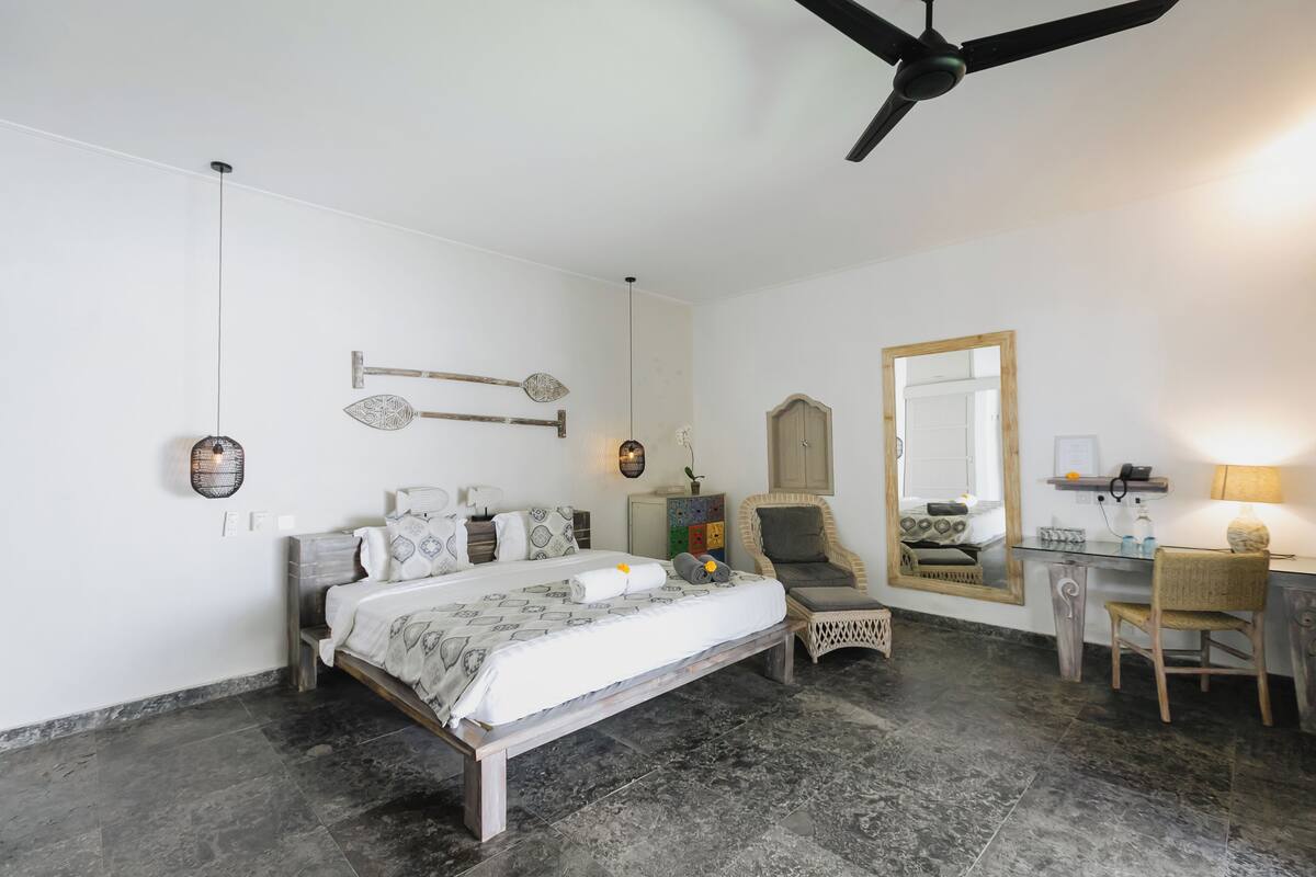 Une chambre à coucher comprenant un lit, une chaise, un bureau, un miroir et un ventilateur de plafond.