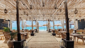 An outdoor dining area with thatched umbrellas, wooden tables, and chairs, overlooking a beach.