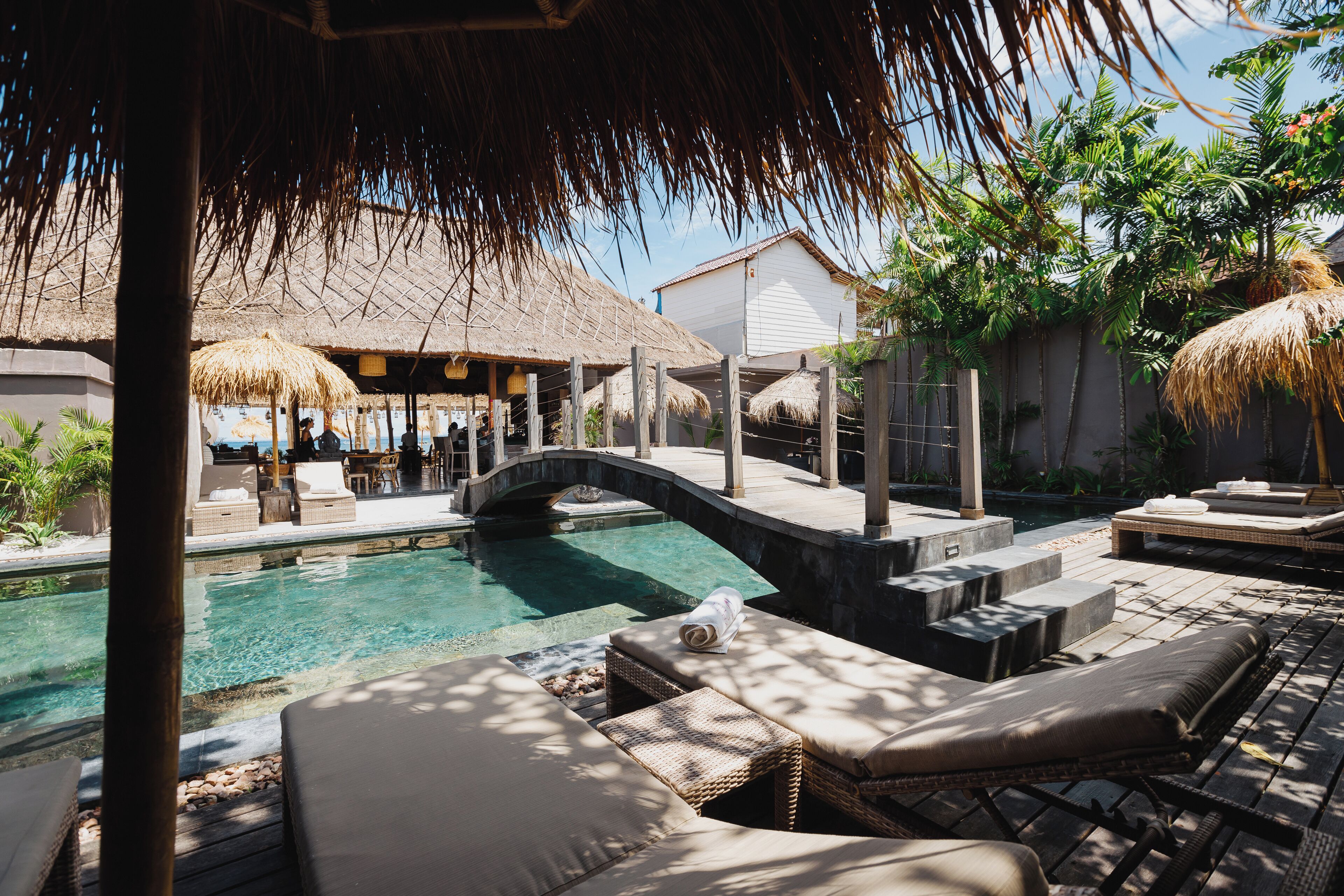 A pool area with lounge chairs, a bridge, and thatched umbrellas.