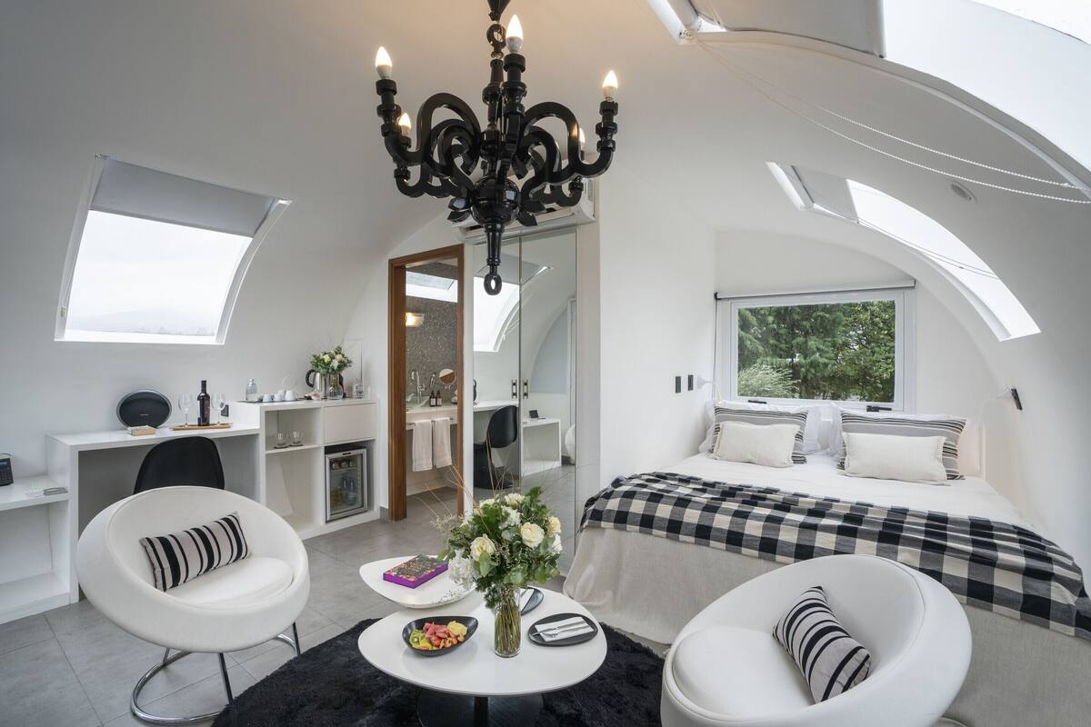 Ein modernes, kleines Schlafzimmer mit Dachfenster, einem Bett mit karierter Bettwäsche, einem Schreibtisch mit Computer und einem Essbereich mit Tisch und Stühlen.