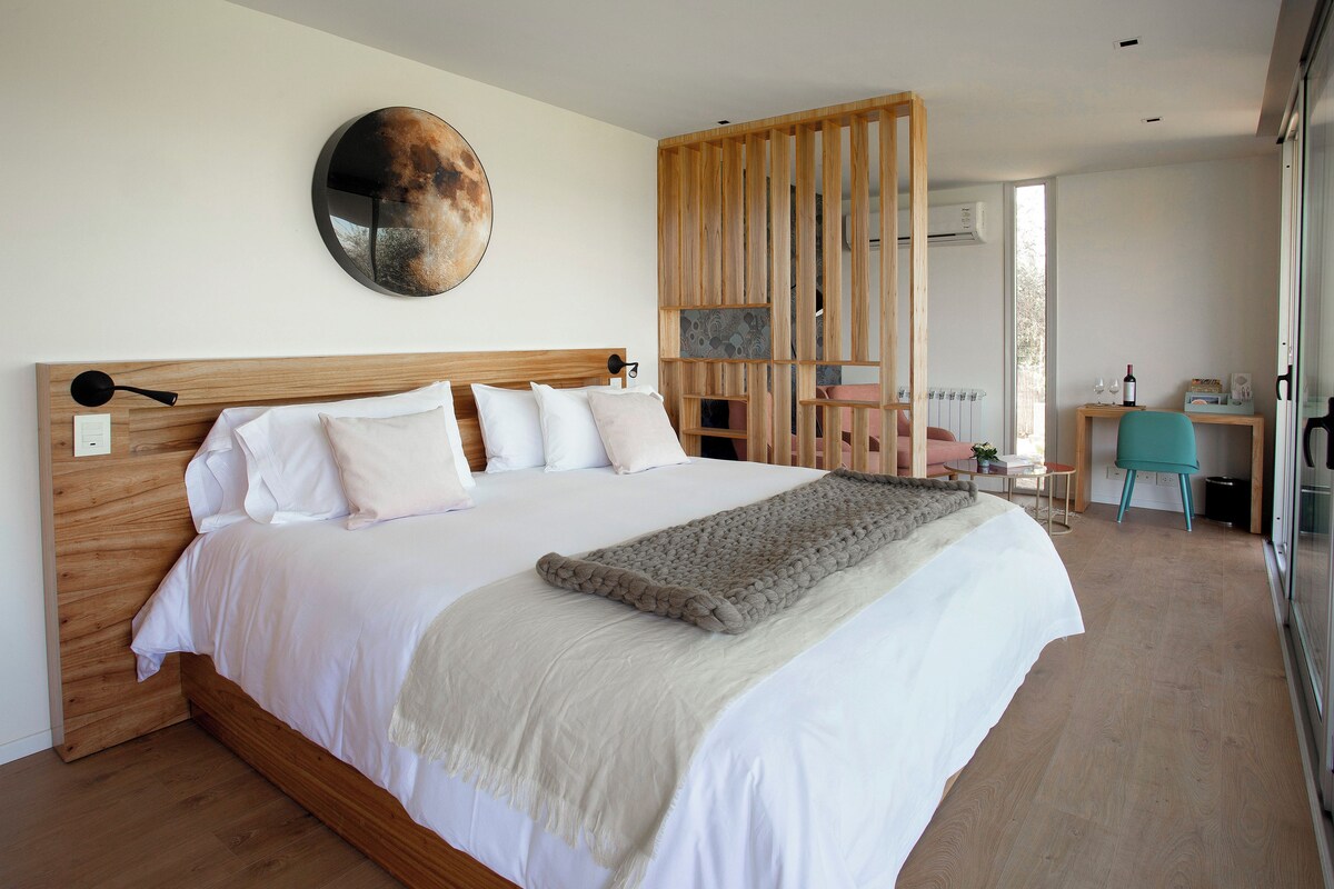 Un dormitorio con cabecera de madera, una cama grande con ropa blanca, un espejo redondo, un separador de madera, un escritorio con una silla y una ventana.