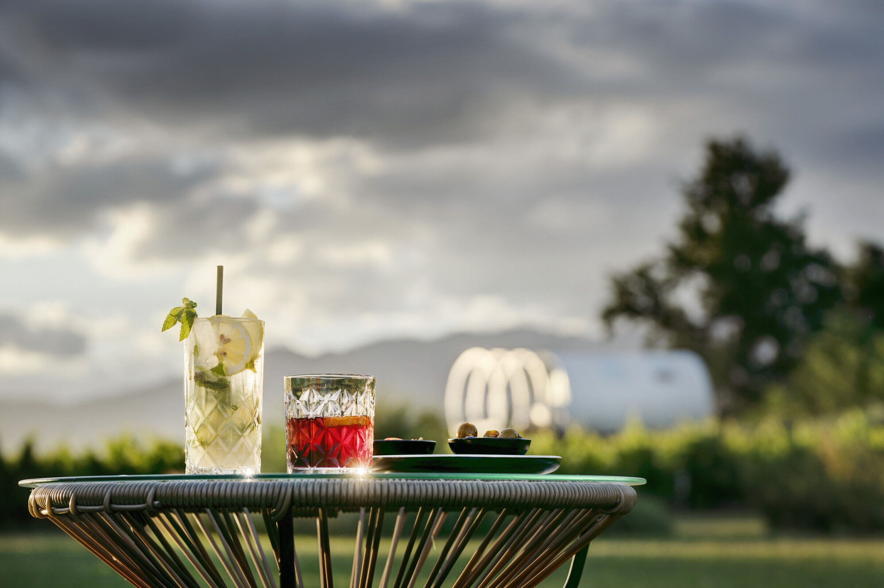 A table with two glasses of beverages, a plate with cookies, and a backdrop of a cloudy sky and mountains.