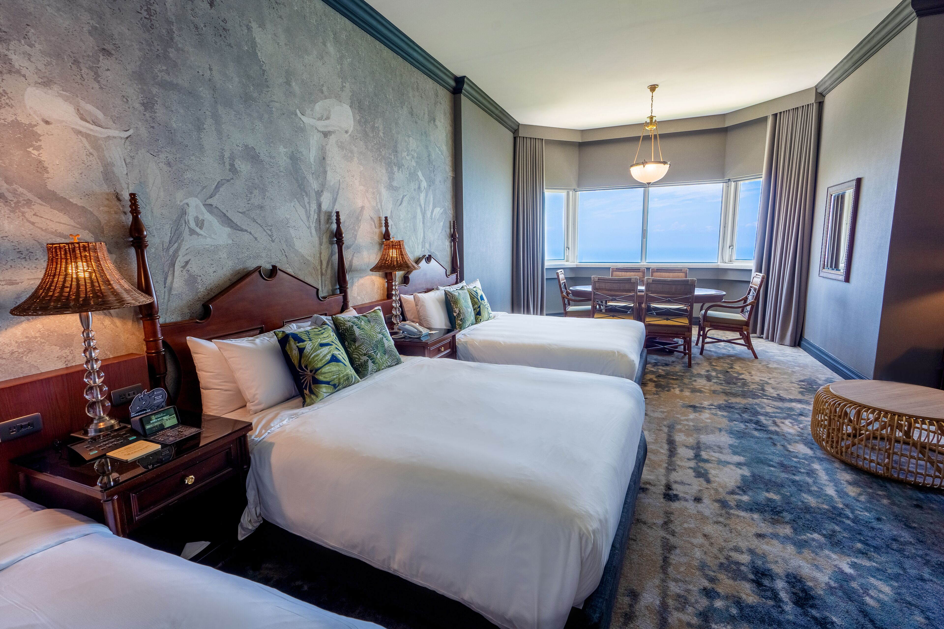 A hotel room with two beds, a dining table, and a view of the sky.