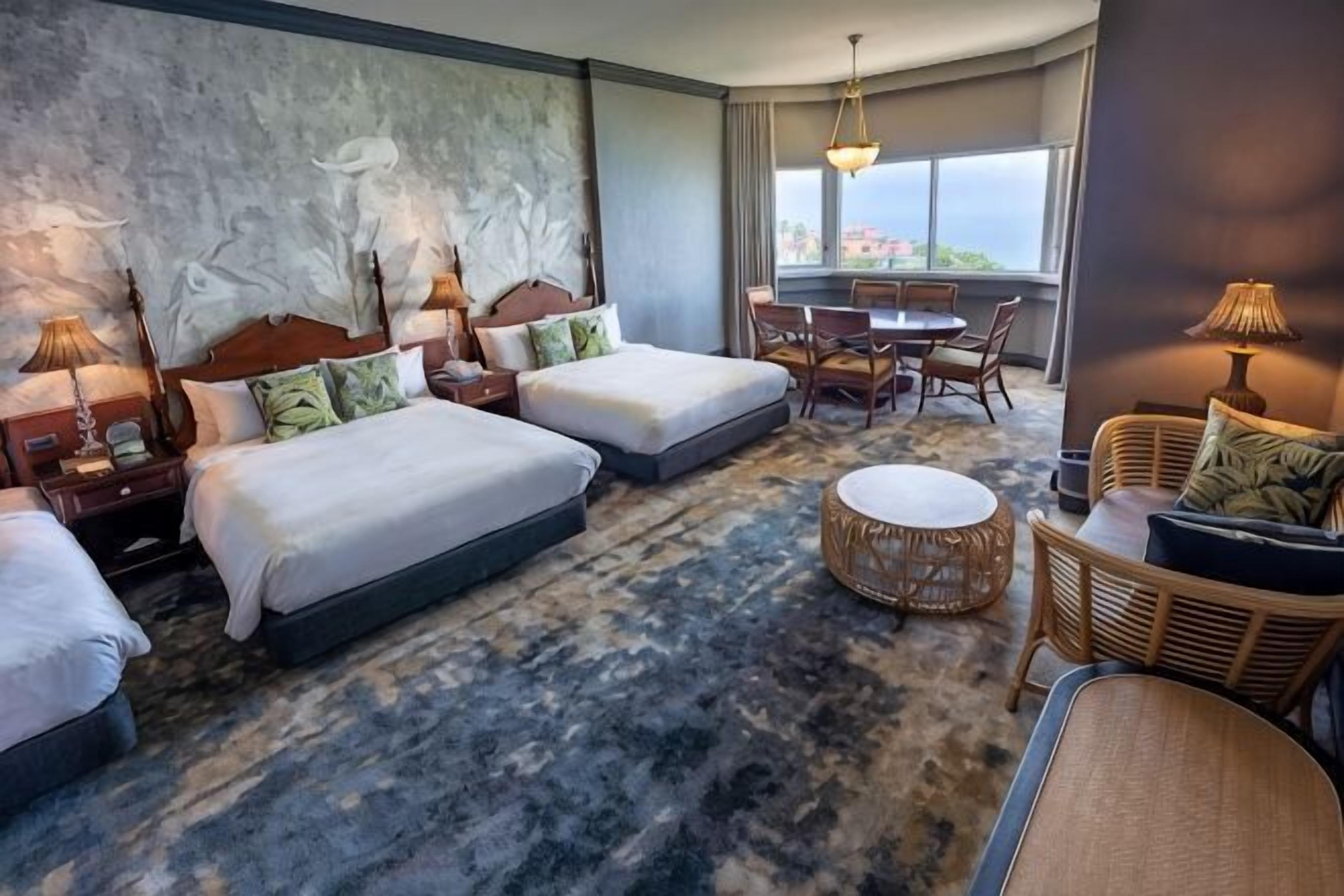 A hotel room with three beds, a dining table, and a view of the city.