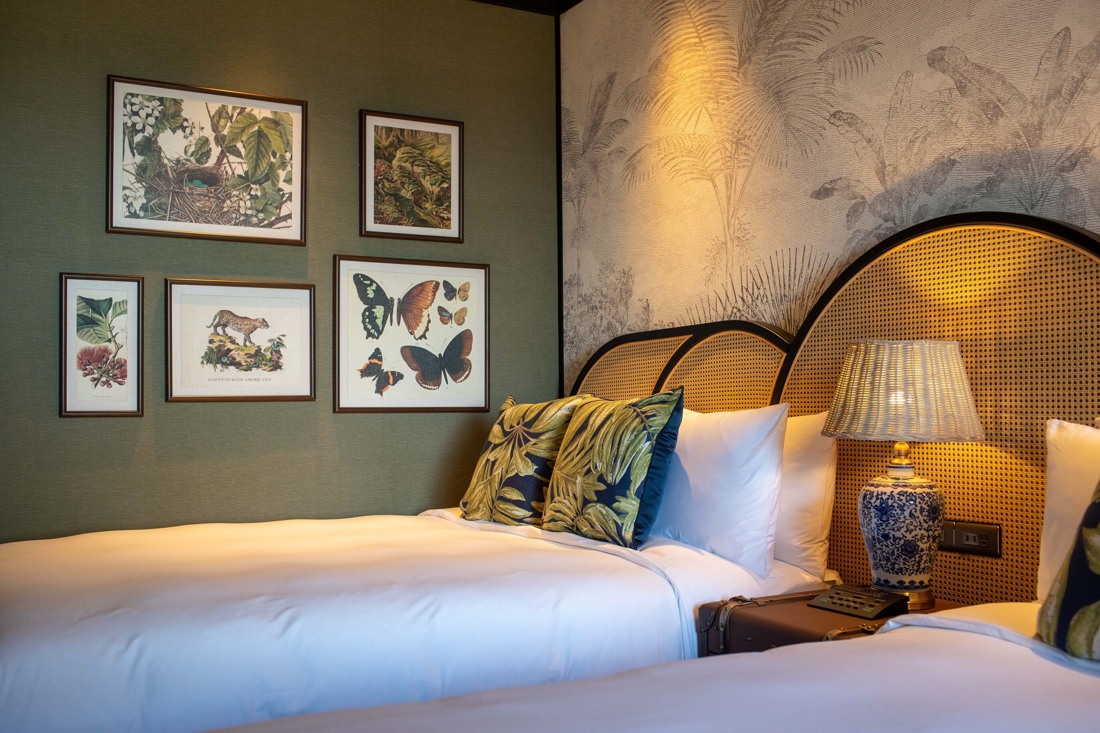 A hotel room with a bed, a nightstand, a lamp, and framed botanical prints on the wall.