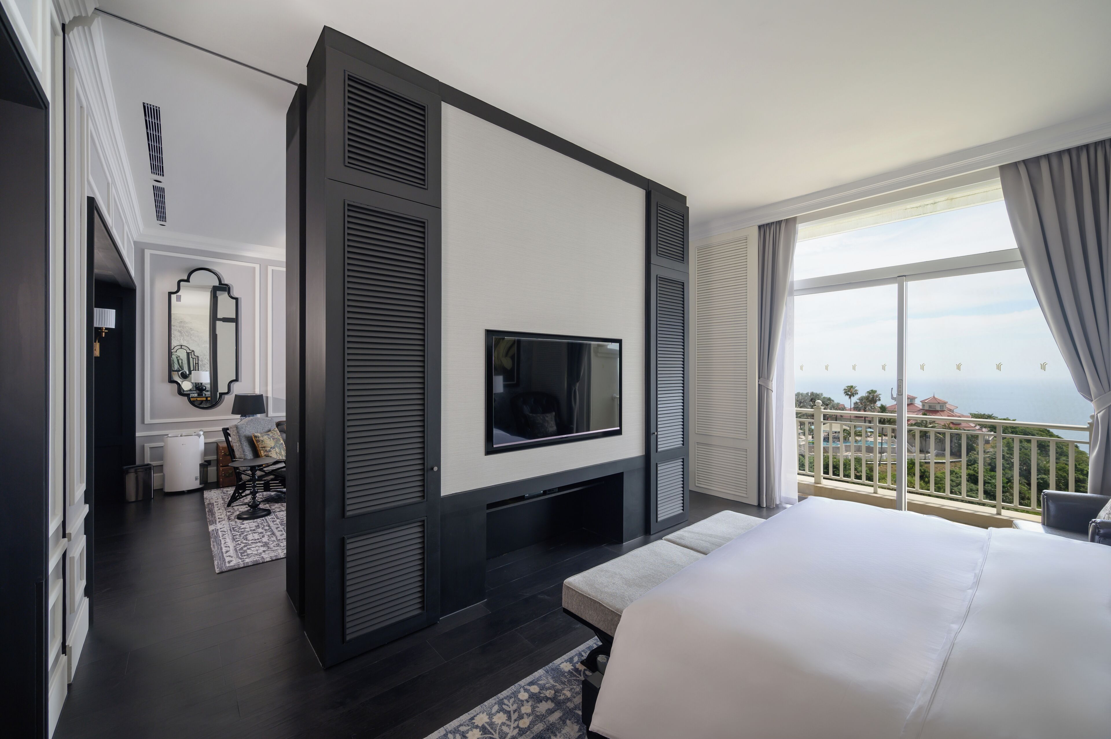 A modern hotel room with a large bed, a TV mounted on the wall, a balcony with a view, and a small seating area with a lamp.