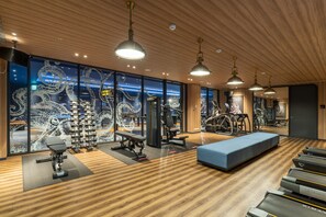 A modern gym with wooden floors, exercise equipment, and large windows with decorative artwork.