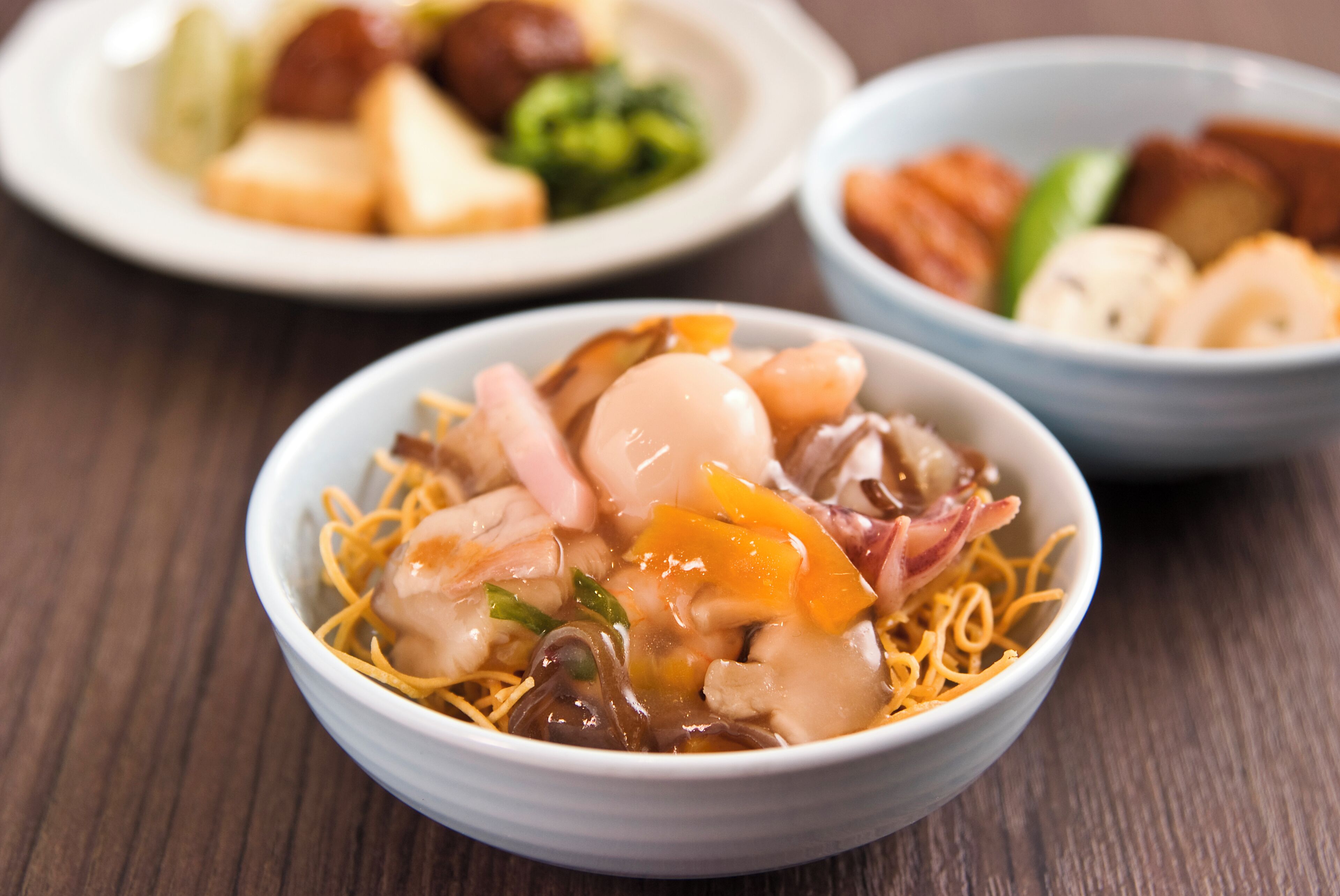 A bowl of noodles with various toppings, a plate with different food items, and a bowl with more food items on a wooden table.