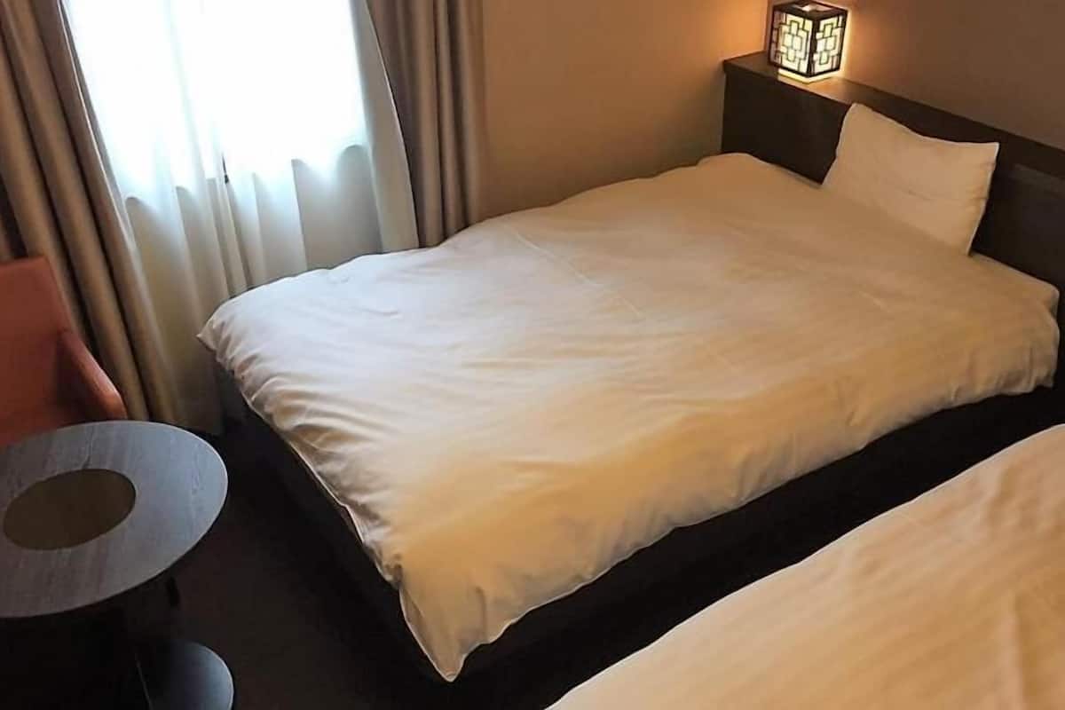A hotel room with a bed, a chair, a small table, and a nightstand with a lamp.