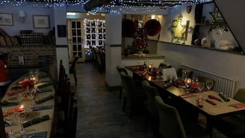 Un restaurant poc il·luminat amb taules preparades per sopar, llums de garlandes penjades i un arbre de Nadal al fons.