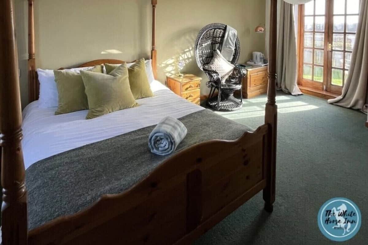 A four-poster bed with a wooden headboard, a rolled towel, and pillows. There is a wicker chair with a cushion, a small wooden nightstand, and a window with curtains.