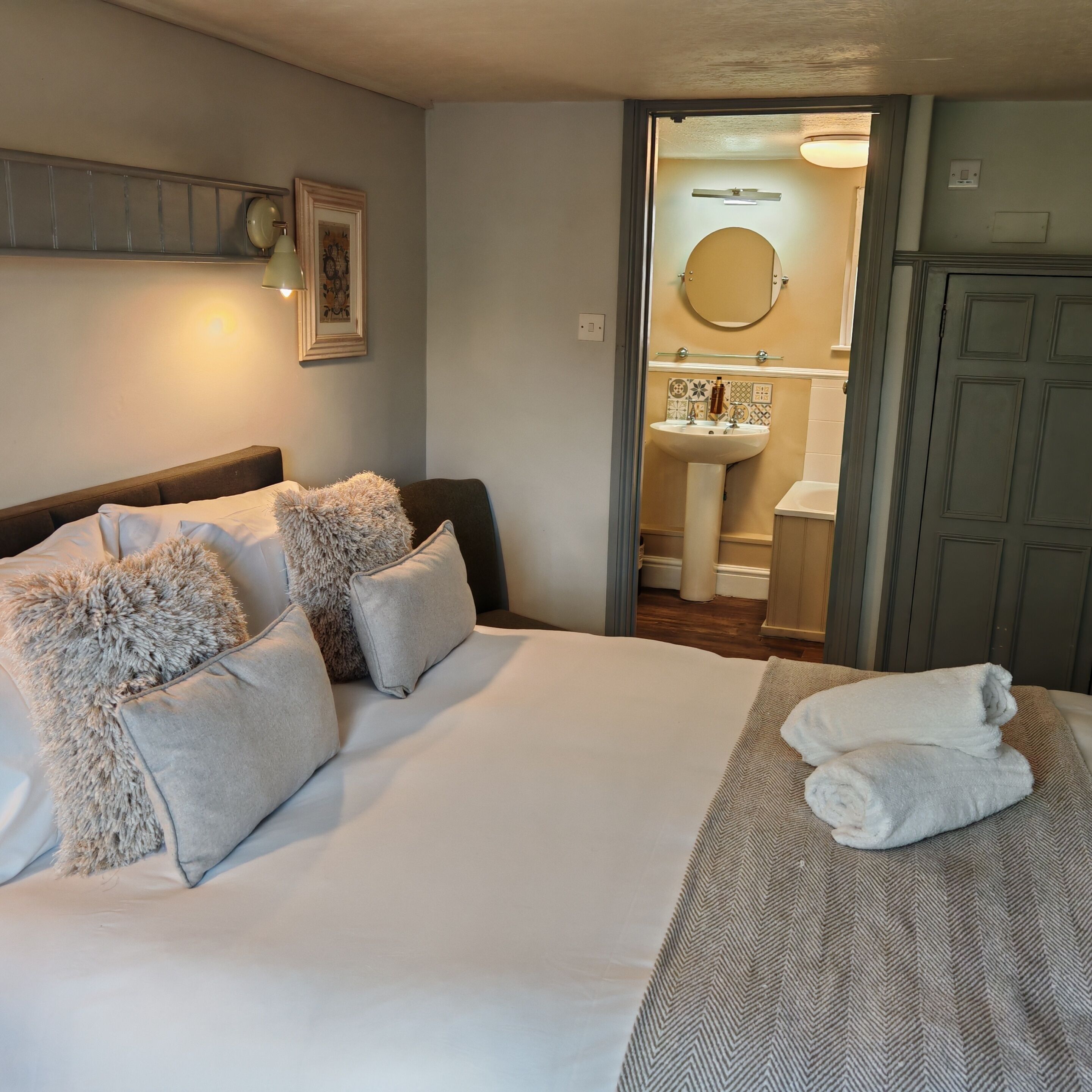 A hotel room with a bed, pillows, a bedside table, a bathroom with a sink and mirror, and a wardrobe.