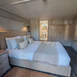 A bedroom with a bed, bedside tables, a nightstand, a dresser, a woven rug, a bathroom with a sink and mirror, and a wardrobe. - The White Horse Inn (Bicester)