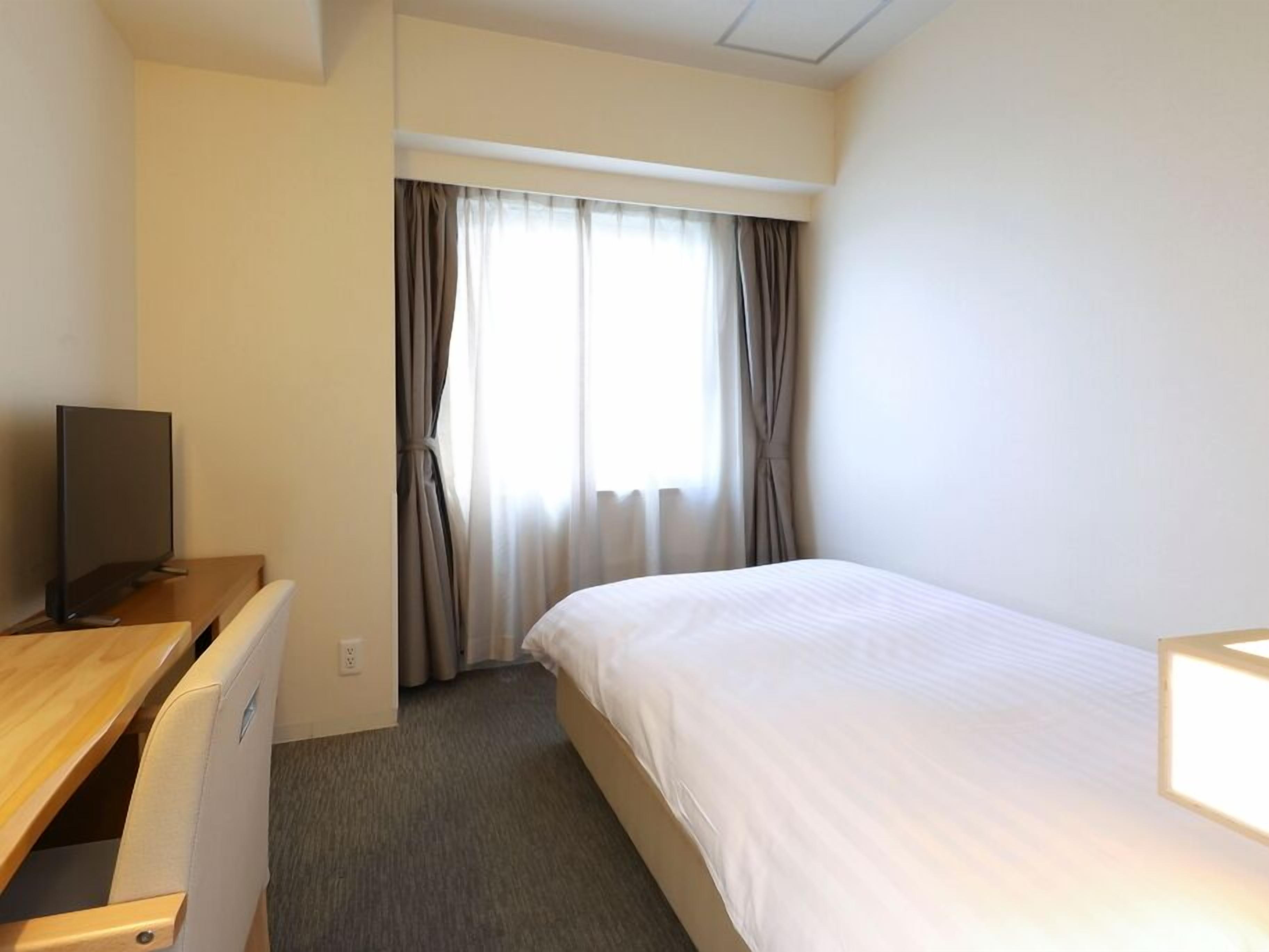 A hotel room with a bed, a desk with a TV, and a window with curtains.