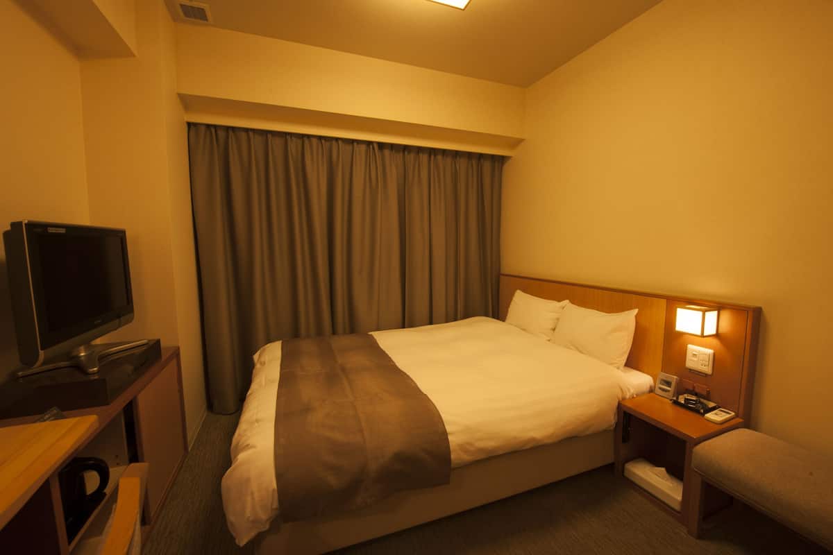 Quarto de hotel com cama, televisão, mesa de cabeceira e cortinas na janela.