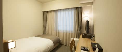 A hotel room with a single bed, a desk with a chair, a desk lamp, a telephone, and a television.