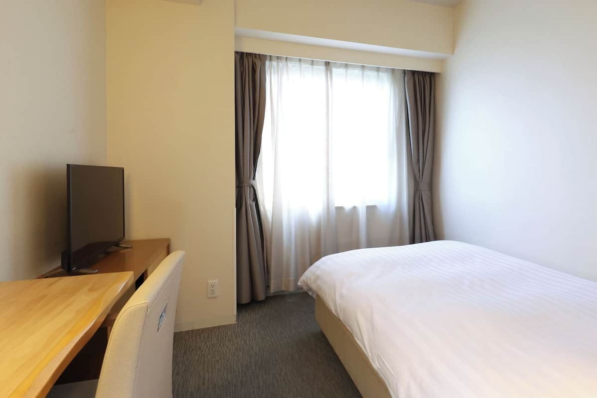 Quarto de hotel com uma cama, uma escrivaninha com televisão, uma janela com cortinas e piso coberto por carpete.