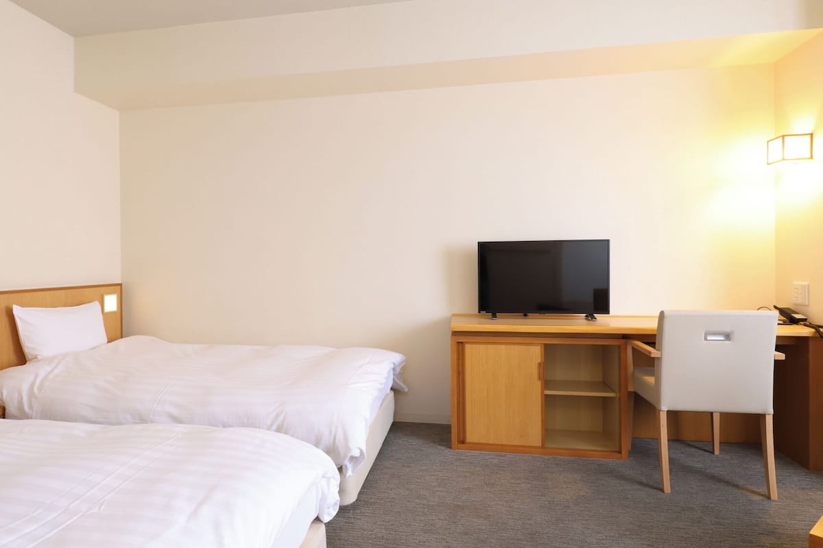 Quarto de hotel com duas camas, uma escrivaninha, uma cadeira, televisão e luminária na parede.