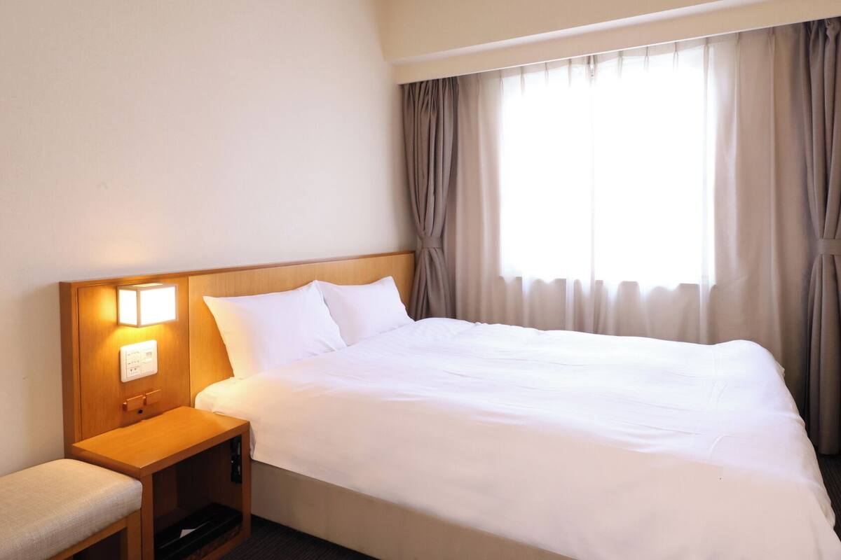 Quarto de hotel com uma cama grande, uma mesinha de cabeceira com abajur, uma cadeira pequena e uma janela com cortinas.