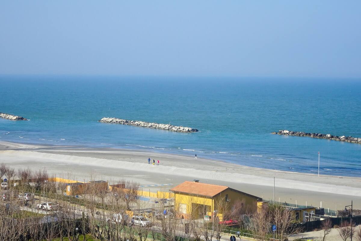 A coastal view with a sandy beach, a rocky breakwater, and a yellow building with a red roof.