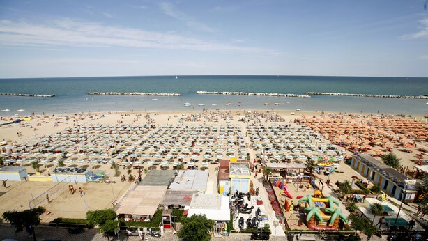 A beach with numerous umbrellas and people. There are boats in the water and a pier in the distance. - Punta Nord Village & Hotel (Rimini)