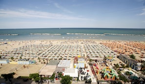 A beach with numerous umbrellas and people. There are boats in the water and a pier in the distance. - Punta Nord Village & Hotel (Rimini)