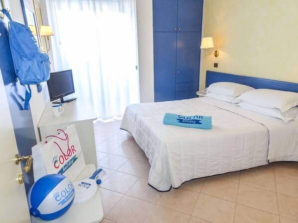 Quarto de hotel com cama, escrivaninha com computador, janela com cortinas e parede azul.