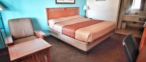 Ein Hotelzimmer mit einem Bett, einem Sessel, einem kleinen Tisch, einem Schreibtisch, einer Lampe und einem durchgegangenes Badezimmertür sichtbaren Bad.