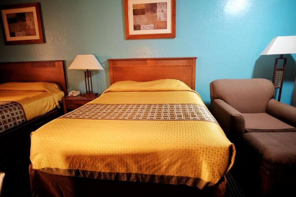 Habitación de hotel con dos camas, una silla y una mesita de noche con lámpara.