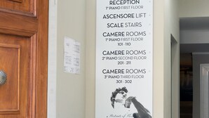 A reception area with a sign listing room types and locations, a potted plant, and a suitcase. - JR Hotels Hermes Firenze (Florence)