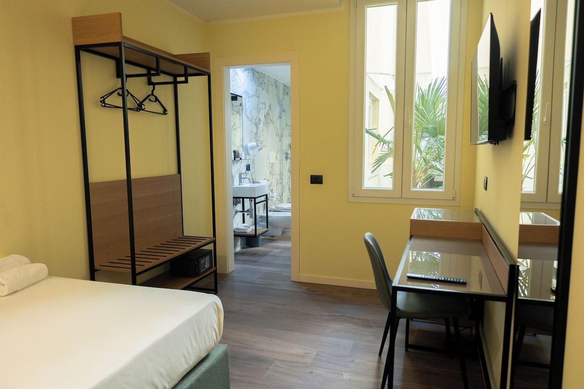 A hotel room with a bed, a desk, a chair, a hanging wardrobe, and a bathroom visible through an open door.