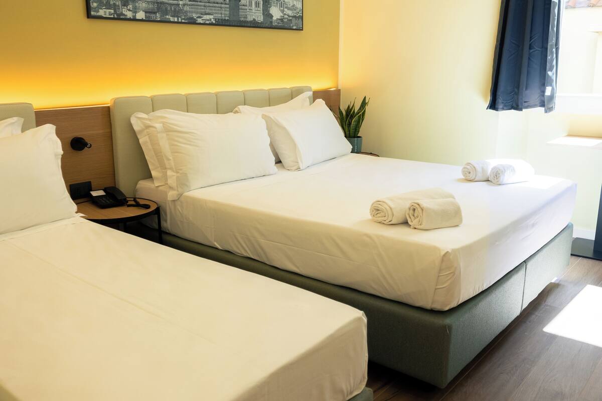 A hotel room with two beds, a bedside table with a phone, a plant, and a window with curtains.
