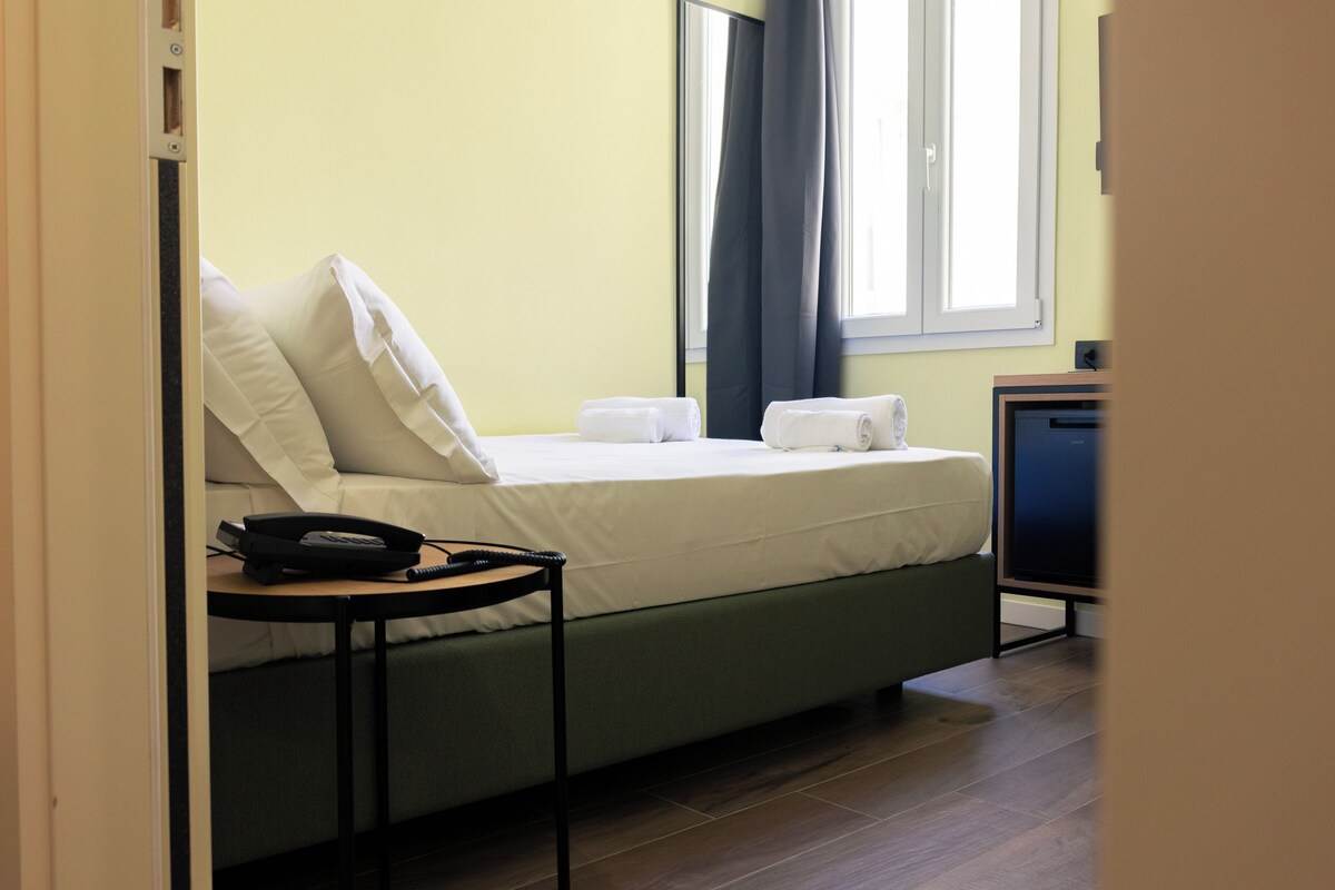 Una cama bien hecha con almohadas y una toalla doblada, una mesita de noche con un teléfono, y un televisor sobre un mueble.