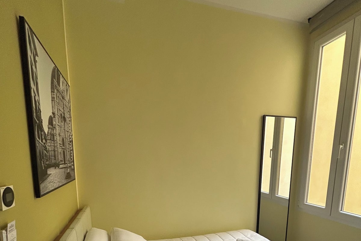 A single bed with white bedding, a bedside table, a wall-mounted mirror, and a window with daylight.