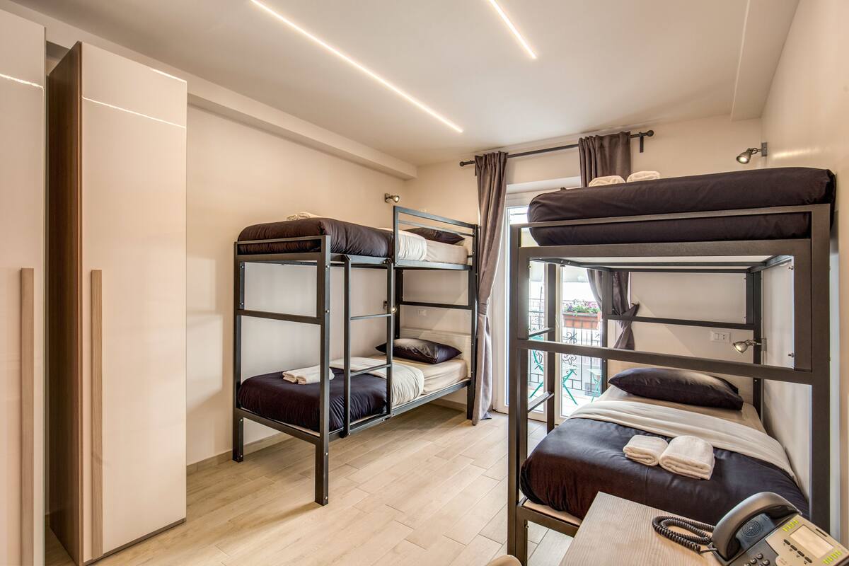 A dormitory-style room with bunk beds, a desk with a telephone, and a window with curtains.