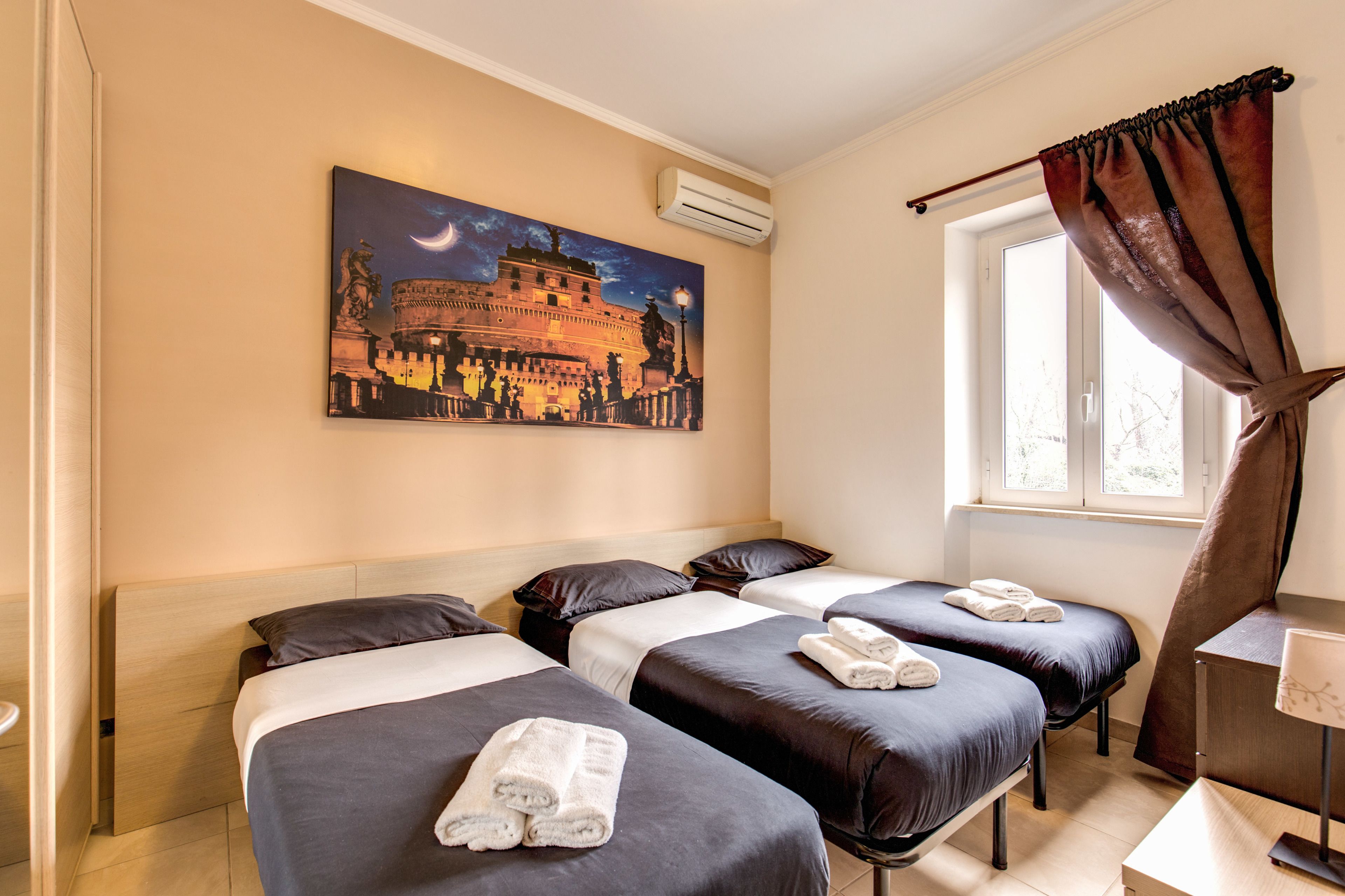 A hotel room with three beds, a painting of a historical building, an air conditioner, a window with curtains, and a bedside table with a lamp.