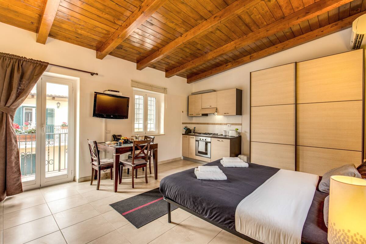 A bedroom with a bed, a desk, a television, and a kitchen area.