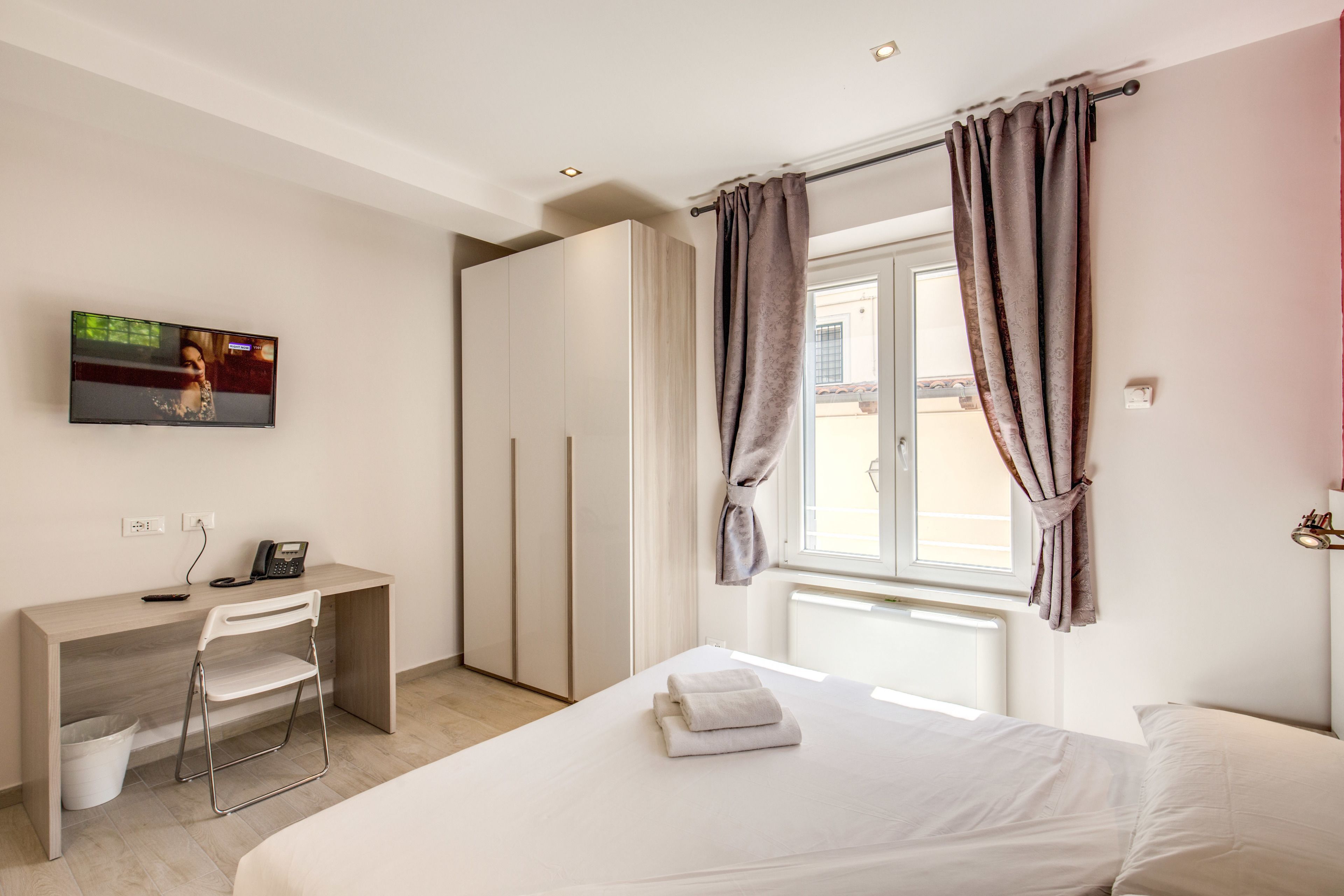 A bedroom with a bed, a desk, a chair, a TV, and a window with curtains.