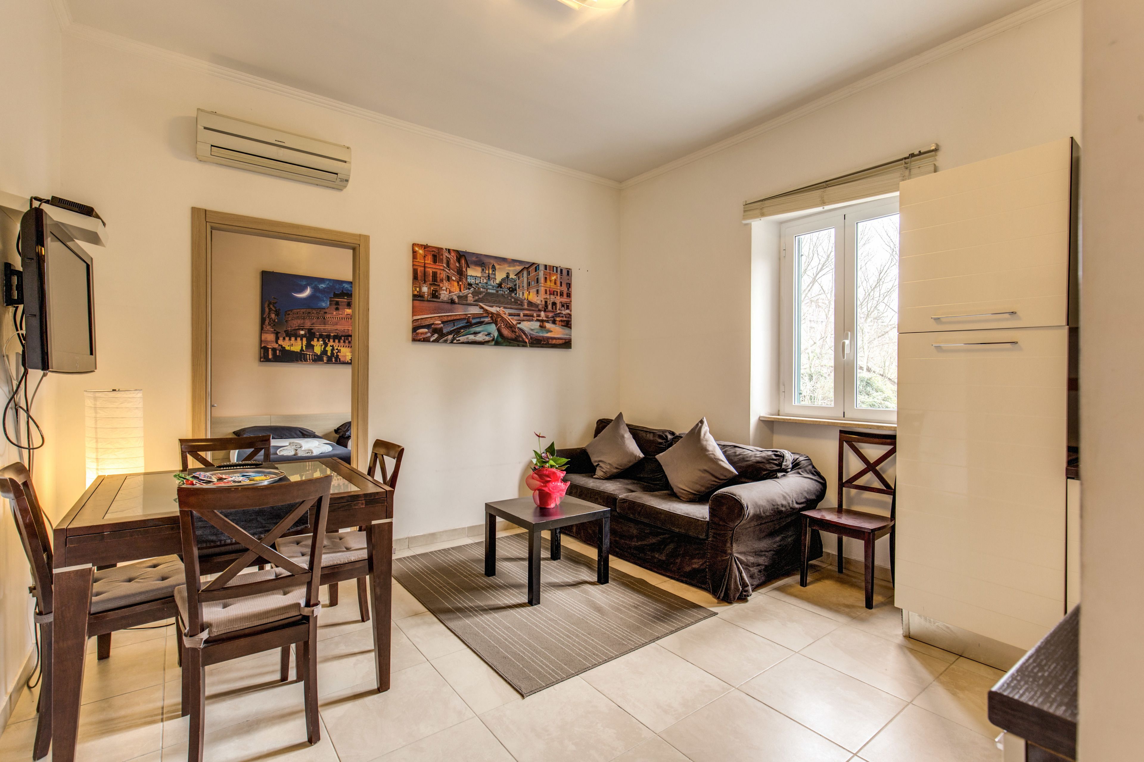A living room with a dining table, chairs, a sofa, and a coffee table. There are two paintings on the wall, a window, and an air conditioning unit.