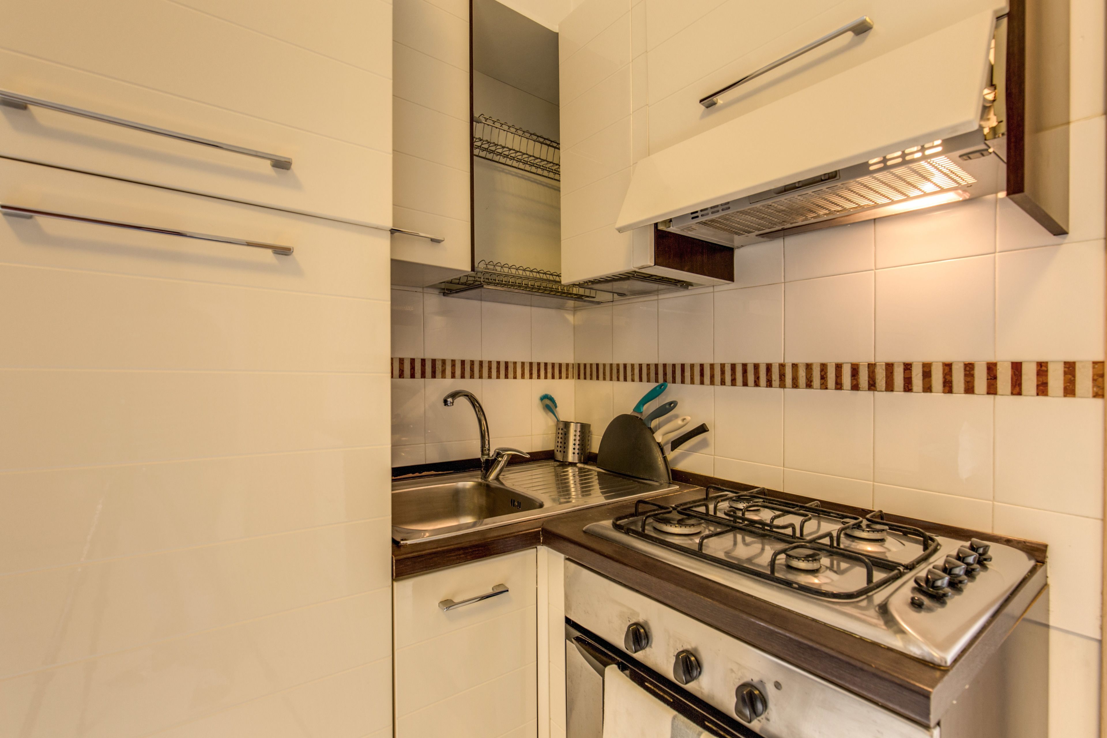 Una cocina con armarios blancos, un lavamanos de acero inoxidable, una estufa a gas y una campana extractora.