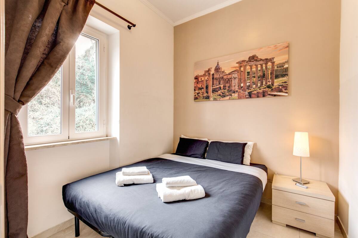 A neatly made bed with folded towels, a bedside lamp, and a painting of ancient ruins on the wall.