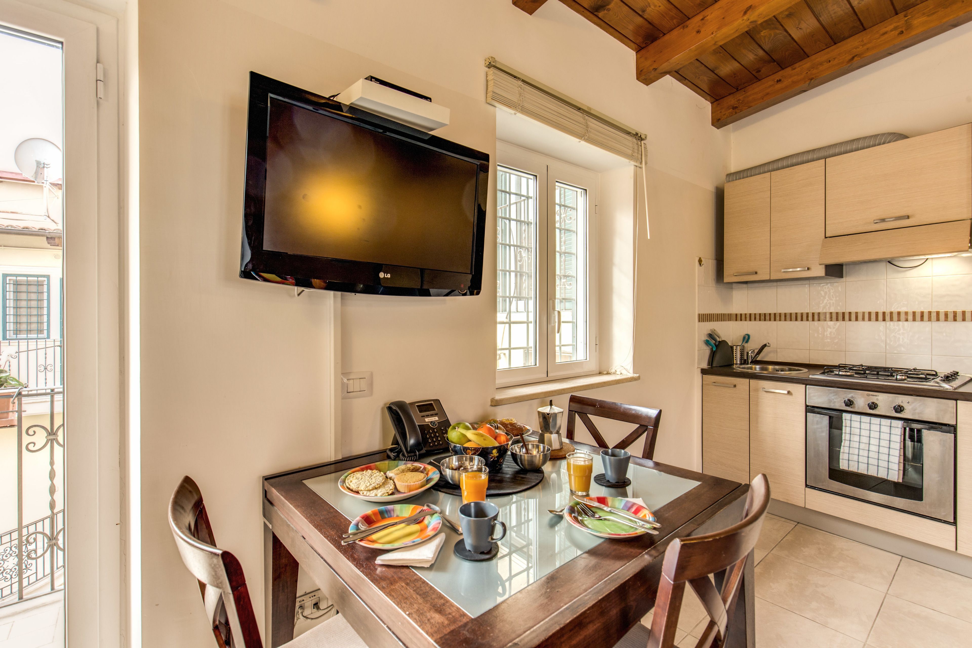 A dining area with a table set for breakfast, a television mounted on the wall, and a kitchen with cabinets and a stove.