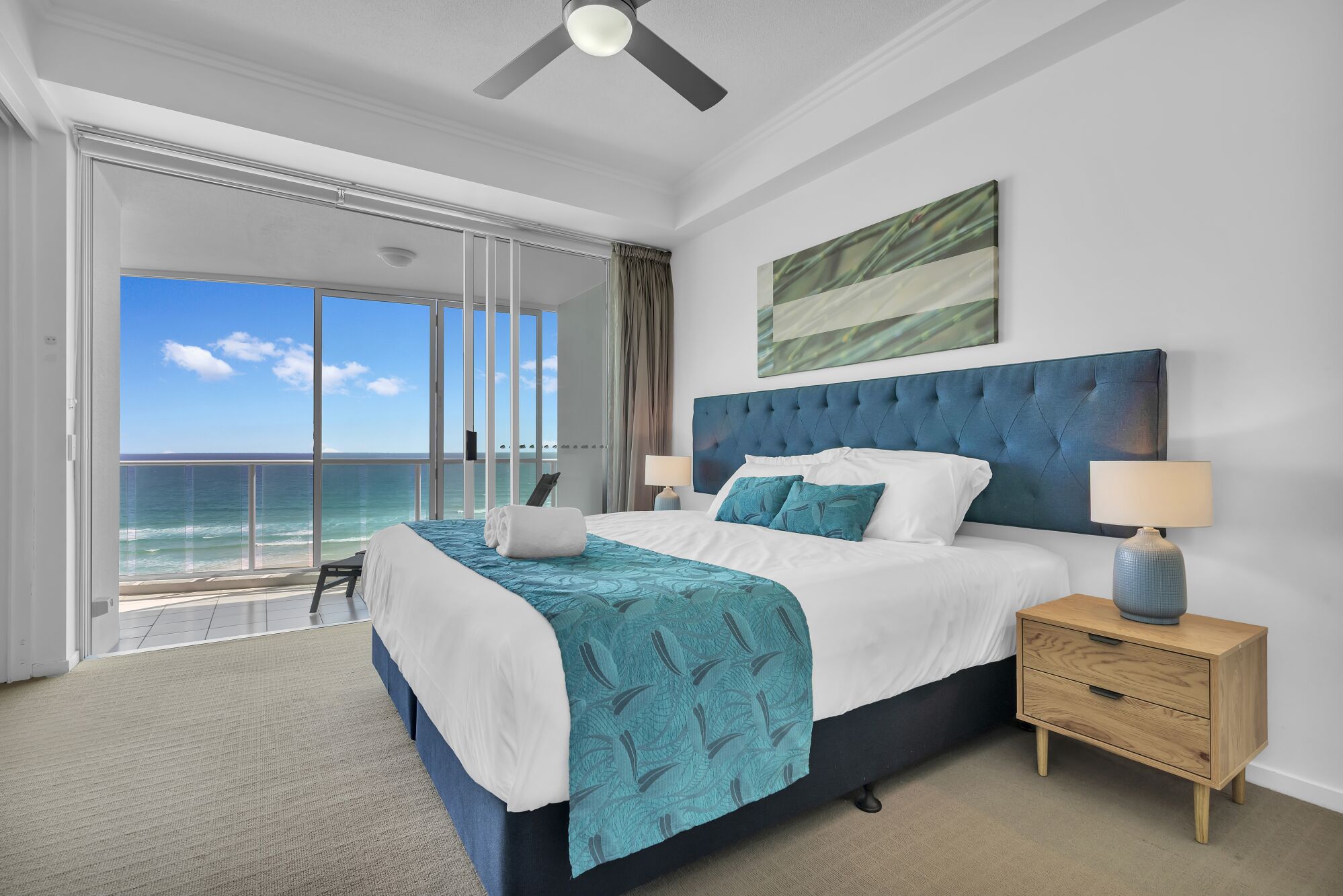 A bedroom with a large bed, a view of the ocean, a ceiling fan, and a nightstand with a lamp.