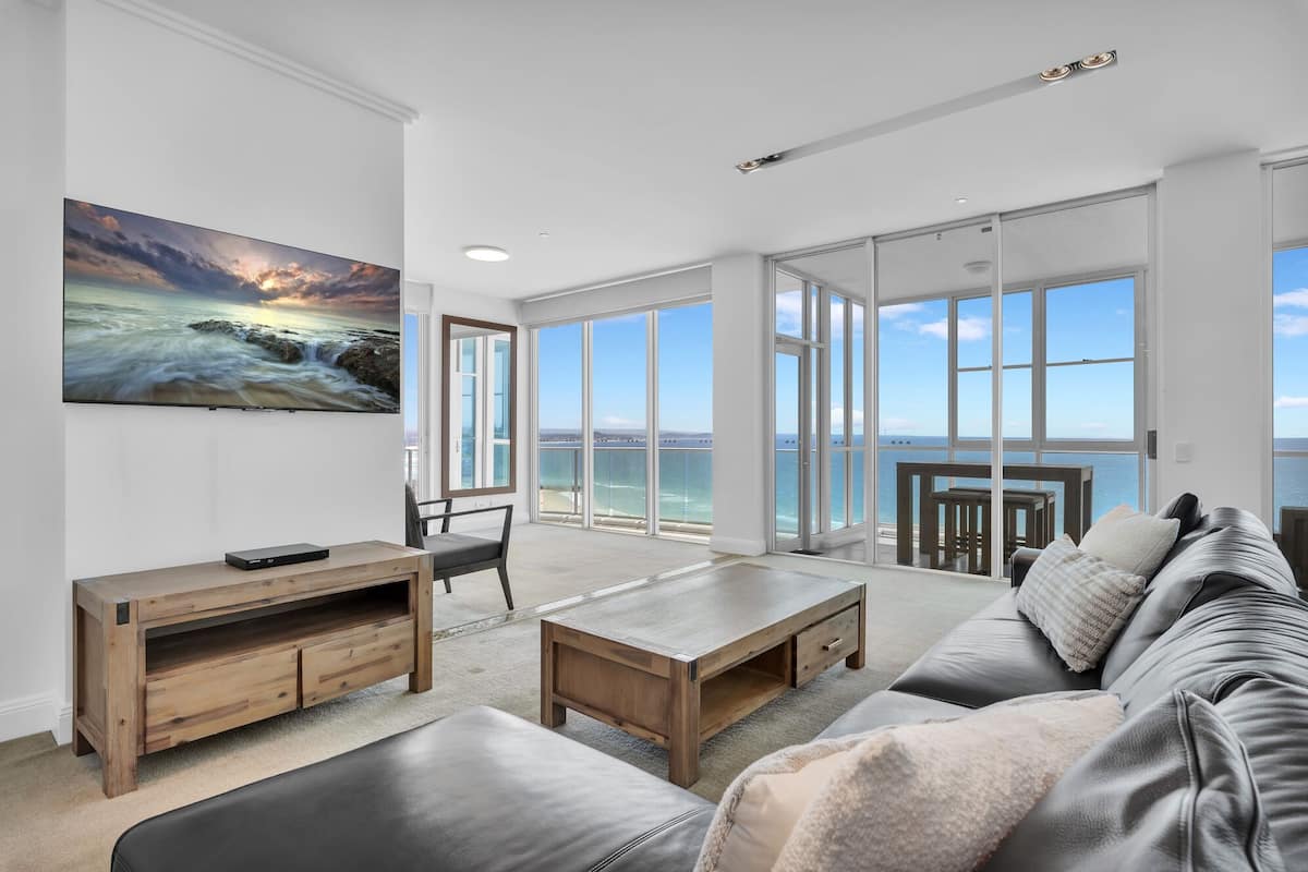 A spacious living room with a large window overlooking the sea, a wooden coffee table, a sofa, and a TV mounted on the wall.