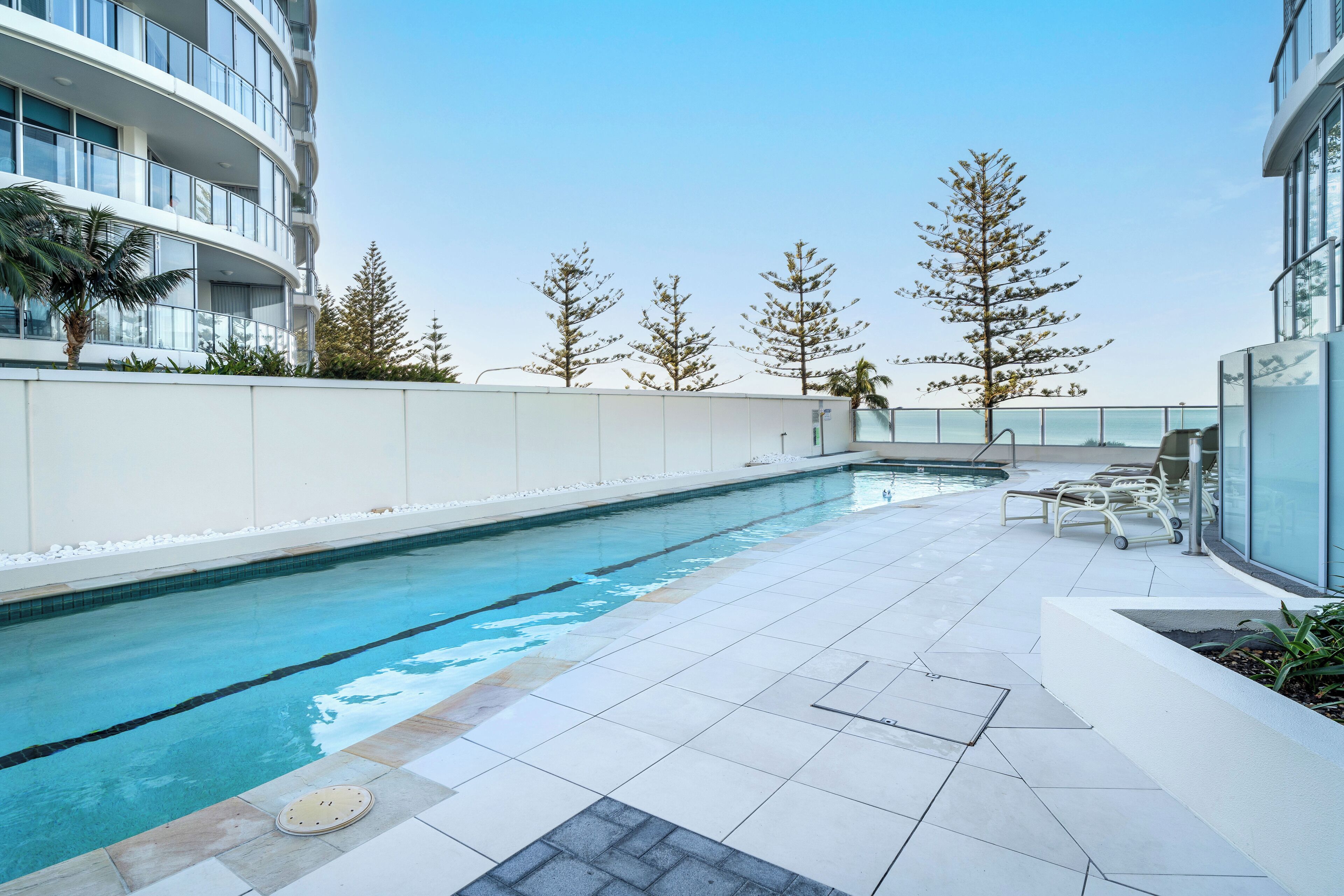 Ein moderner Außenpool mit klarem, blauem Wasser, umgeben von einer weißen Fliesenveranda und einer niedrigen weißen Mauer.