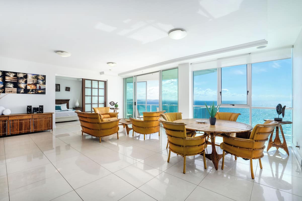 A spacious living room with a large dining table, yellow chairs, and a view of the ocean.