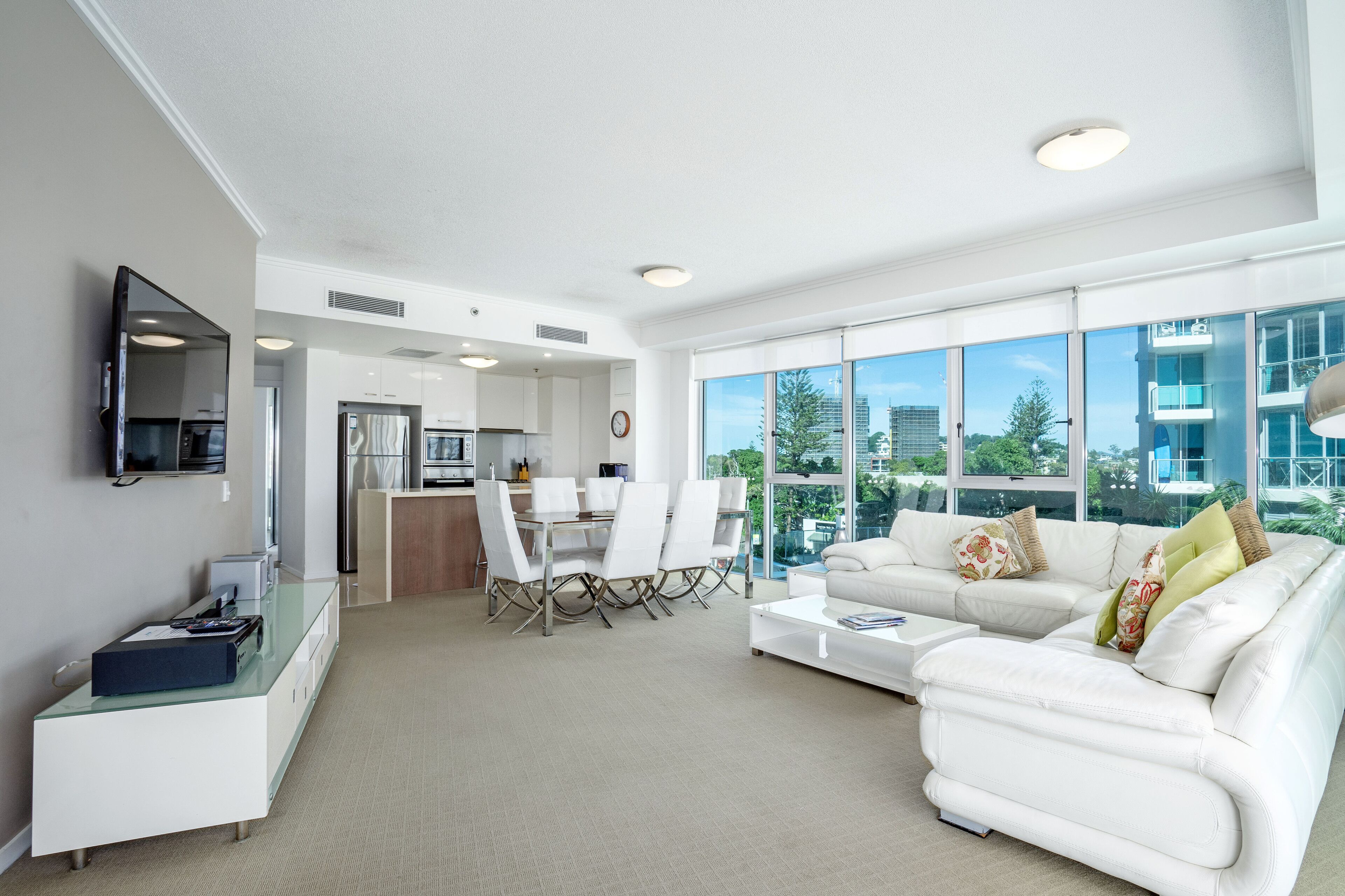 A modern living room with a white sofa, a dining area with a glass table, and large windows offering a view of the city.