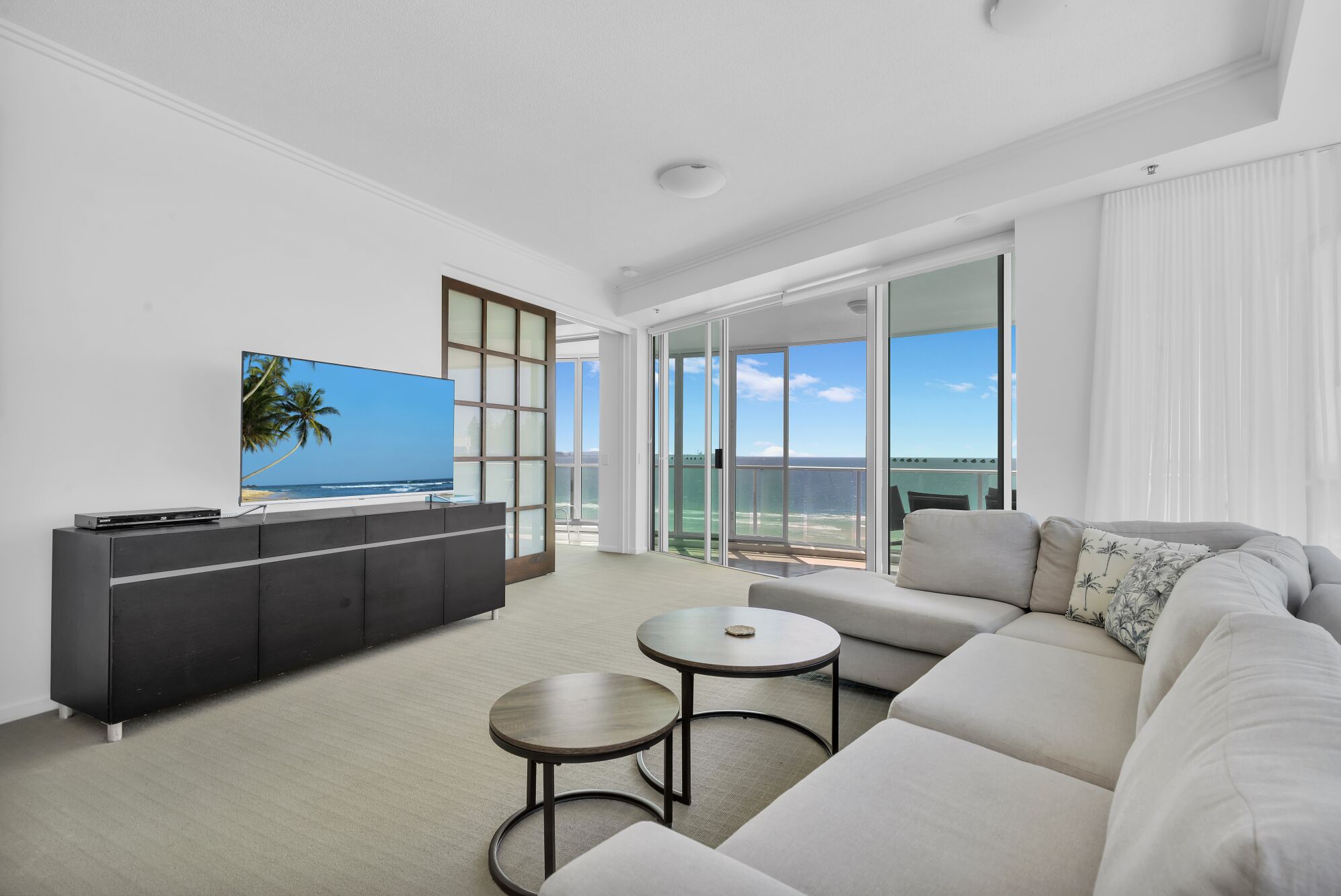 A modern living room with a large flat-screen TV, a sofa, a coffee table, and a sliding glass door leading to a balcony with a view of the ocean.
