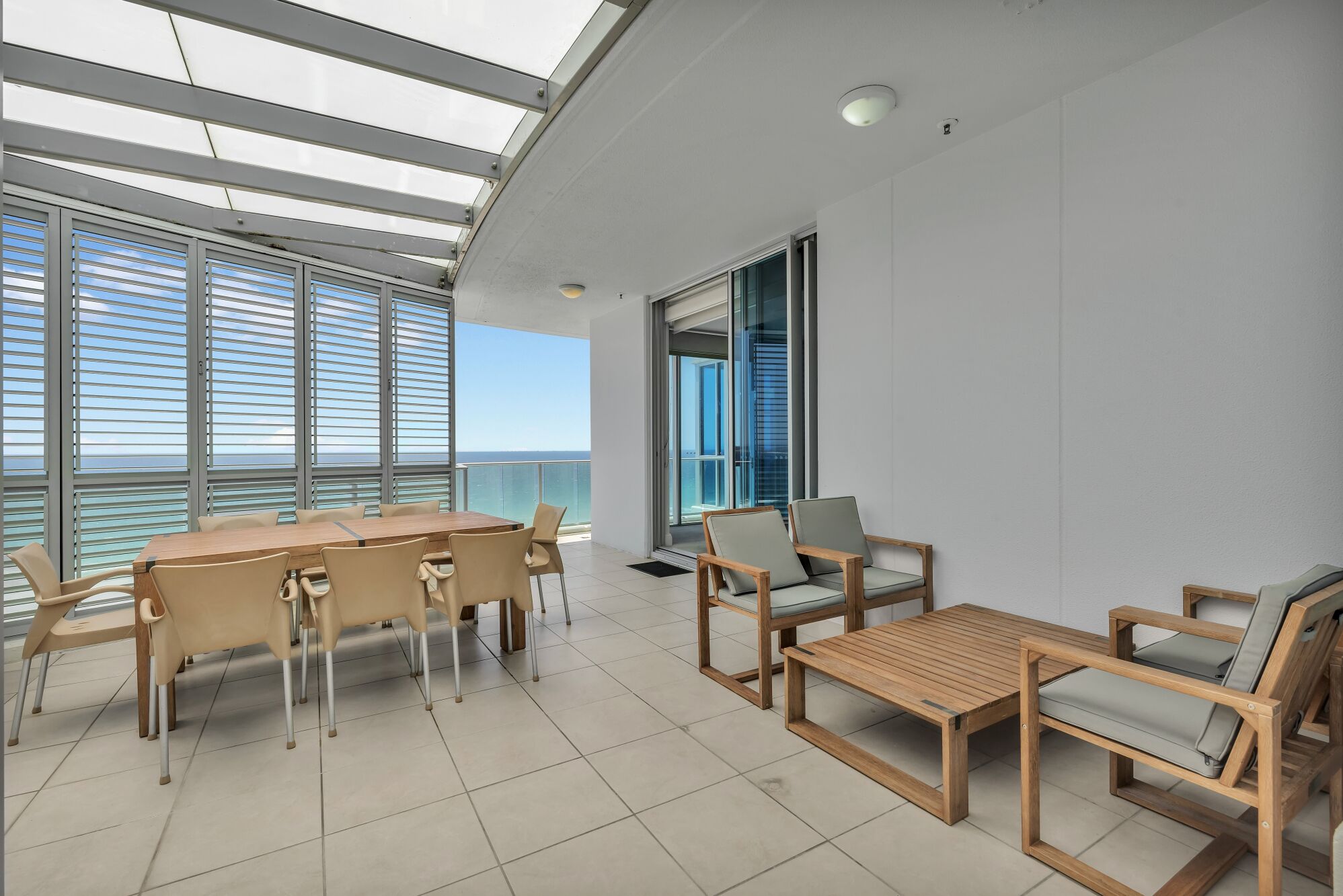 Un balcón moderno con una mesa y sillas de madera, ventanas grandes con persianas y vista al mar.